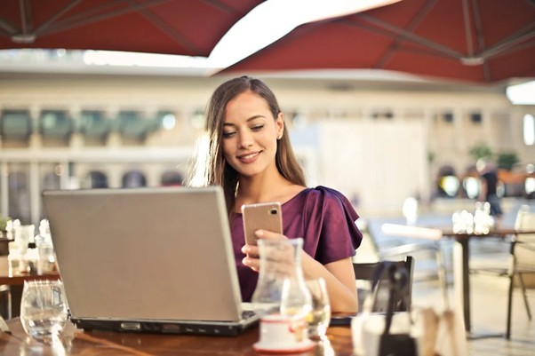 An image of a woman sitting in a cafe looking at her phone and her laptop.